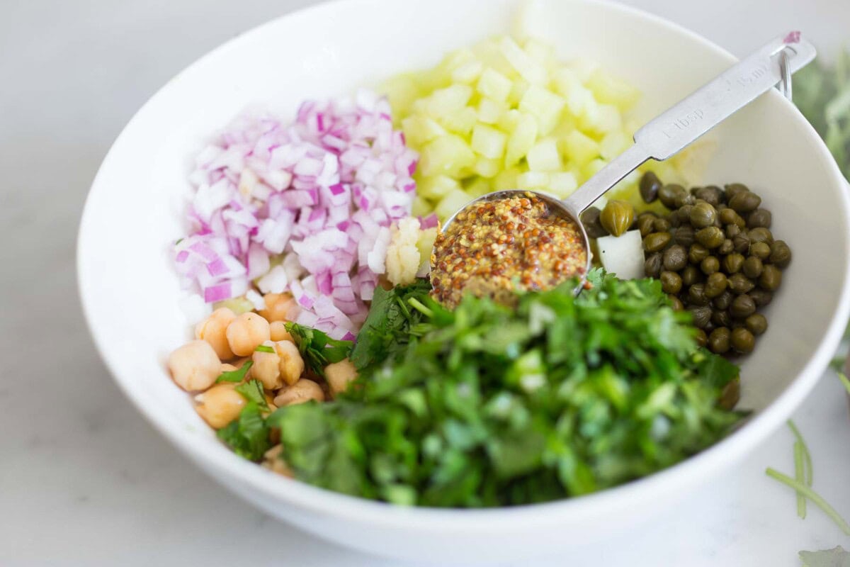 bowl with ingredients for chickpea salad- minced celery and red onion, capers, herbs, chickpeas, and tablespoon of stone ground mustard. 