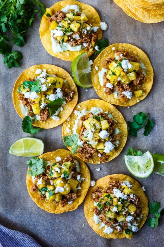 chipotle tostadas with avocado cream, savory ground meat, pineapple salsa, fresh cilantro, and cotija, served alongside lime wedges.