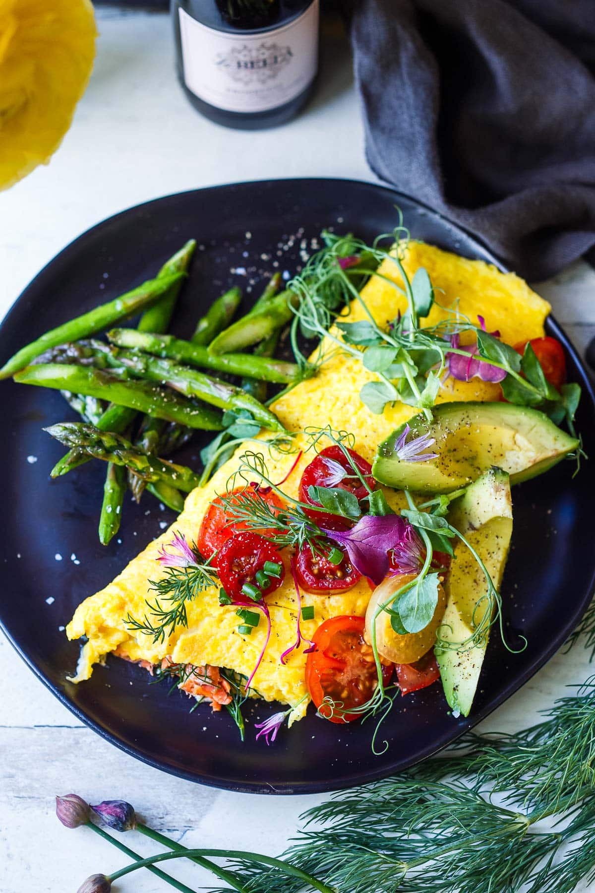 champagne omelette on plate with asparagus, topped with avocado slices, cherry tomatoes, micro greens, and herbs. 