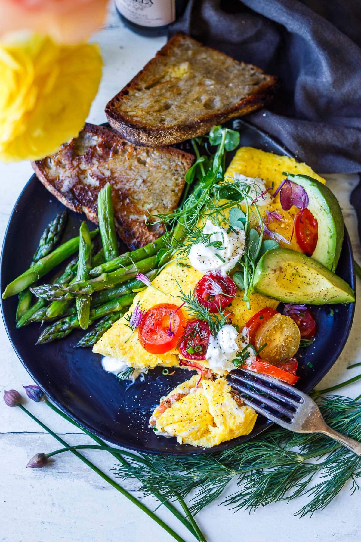 champagne omelette with cherry tomatoes, avocado, micro greens, and creme fresh on top, served on plate with asparagus and toast.