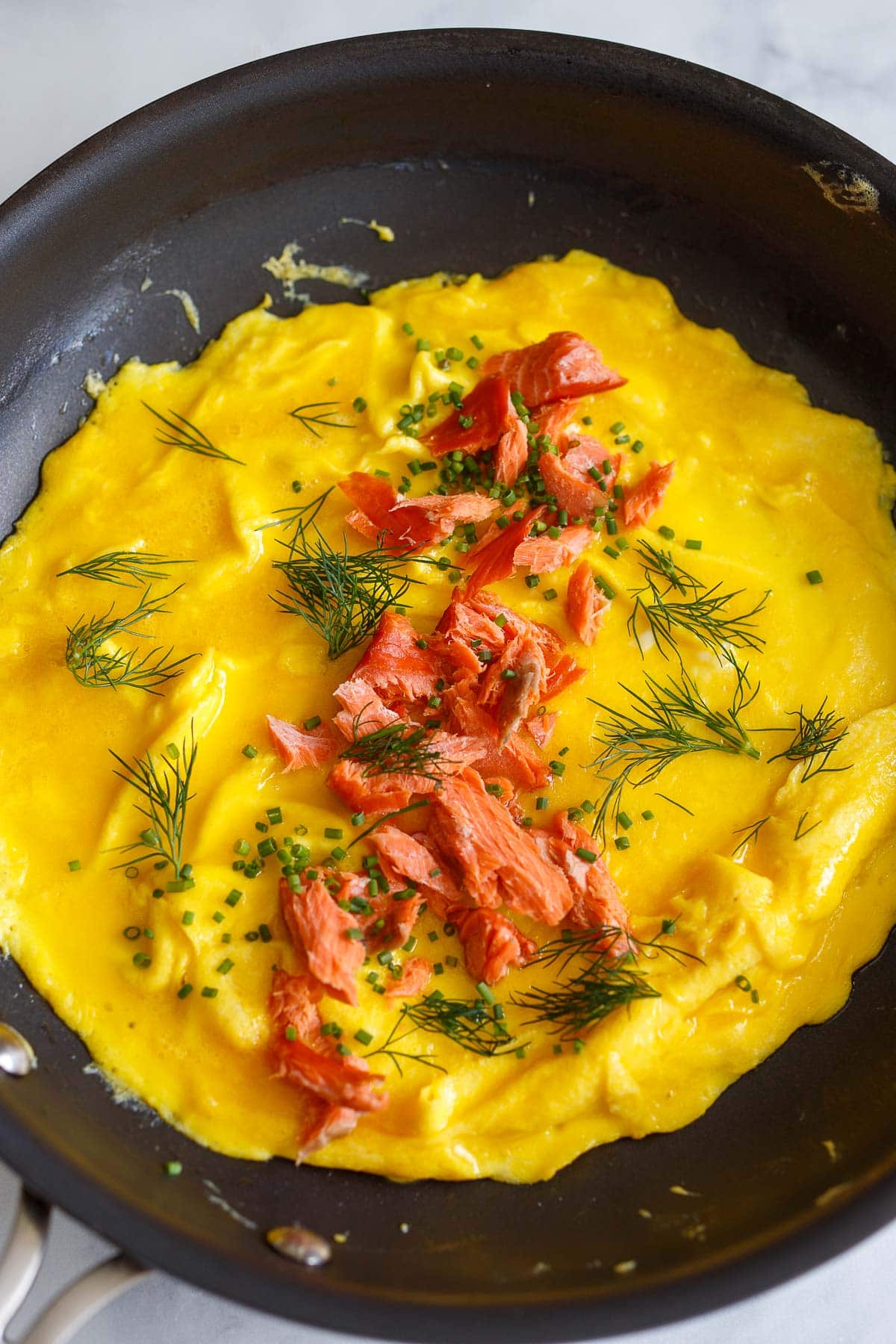 omelette in skillet with flaked salmon and herbs sprinkled over center.