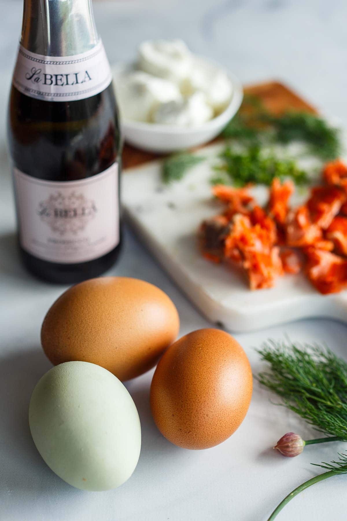ingredients for champagne omelette- 3 farm fresh eggs, multi colored, champagne, and serving board with flaked salmon and fresh herbs.