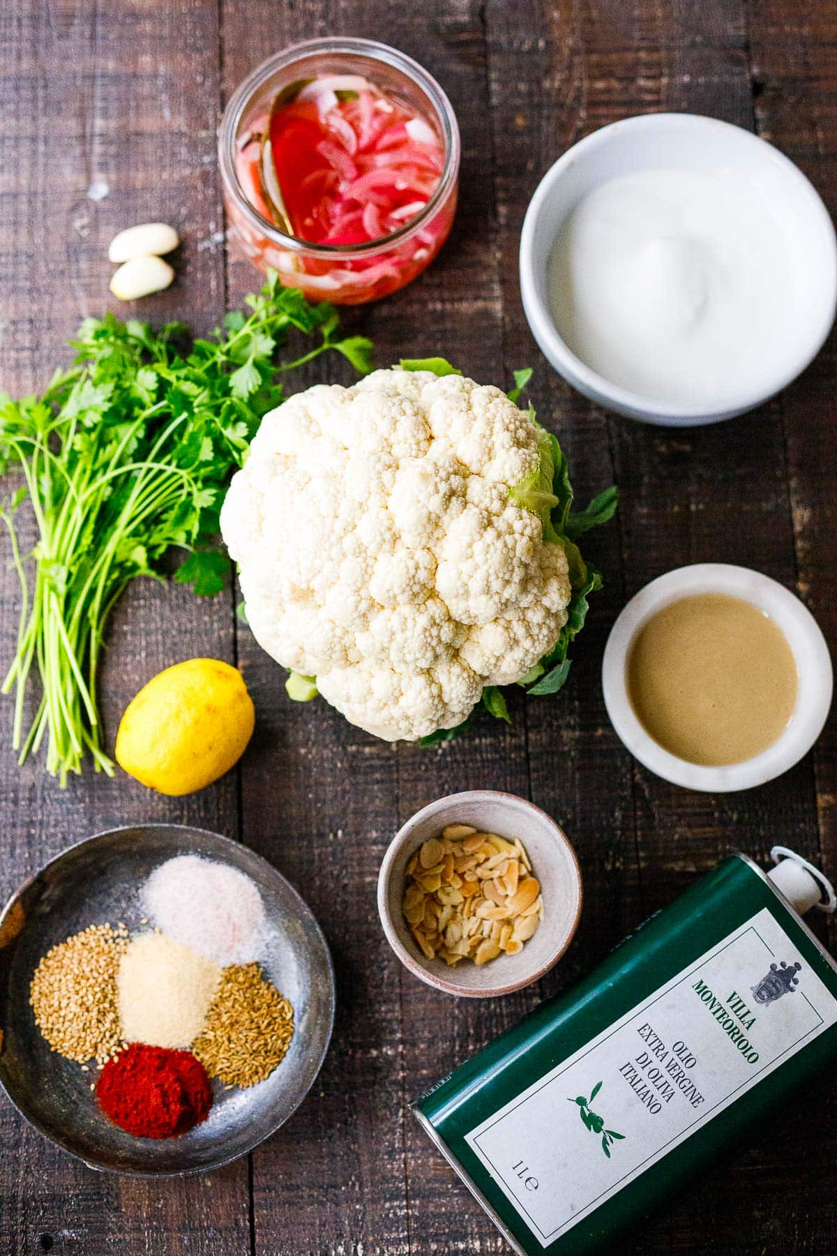 ingredients for crispy cauliflower laid out on wood surface - cauliflower head, yogurt, tahini, pickled onions, garlic cloves, parsley, lemon, almonds, spices, oil. 