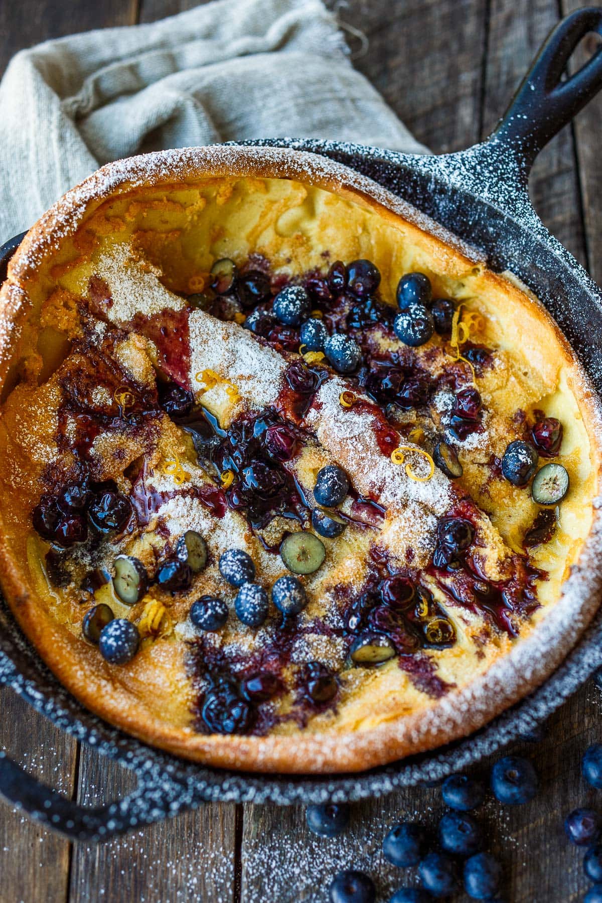 blueberry dutch baby in cast iron skillet, topped with blueberries, blueberry maple syrup, powdered sugar, and lemon zest.